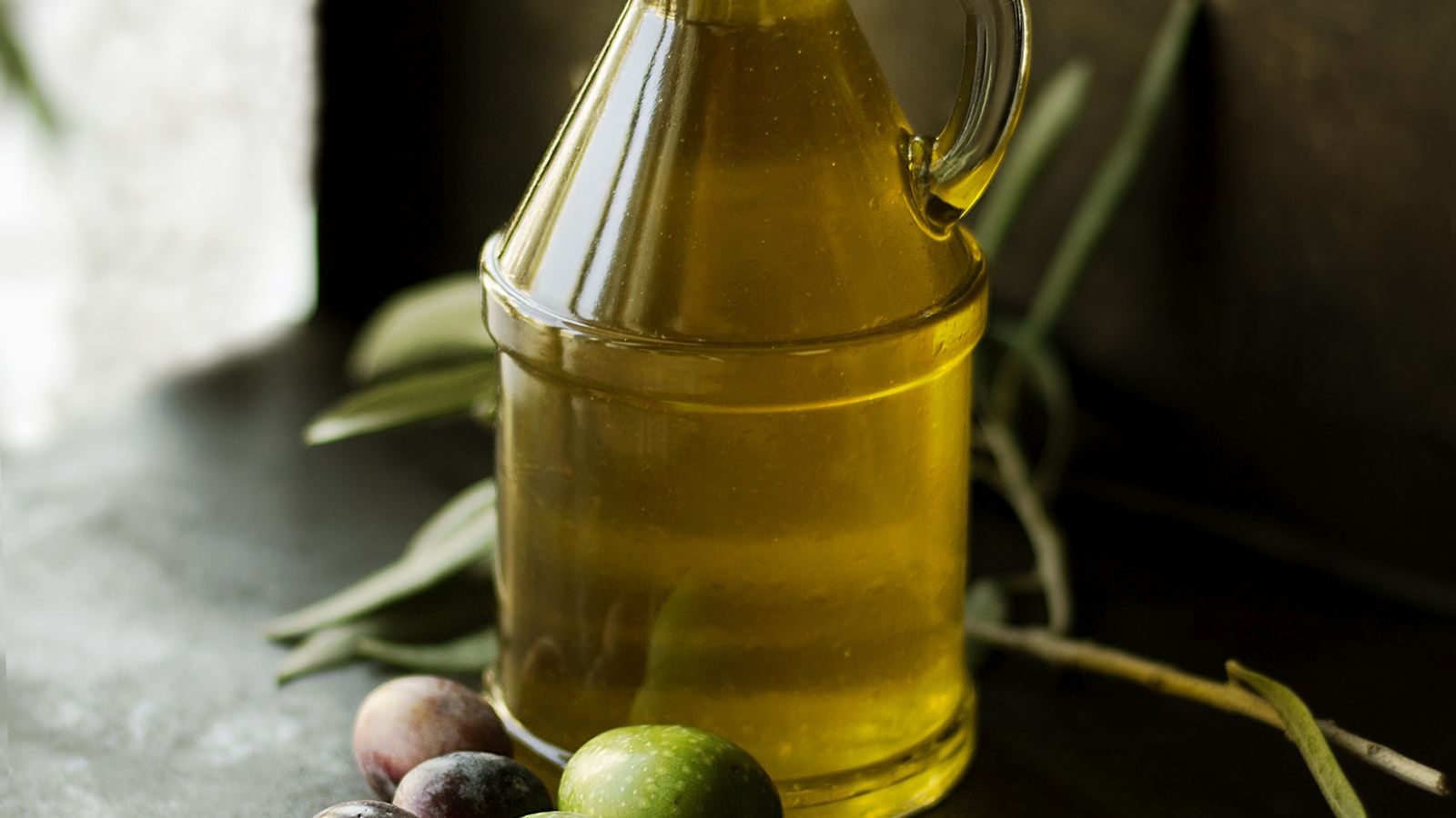 A glass jug of golden olive oil beside a few fresh green and purple olives on a dark surface.