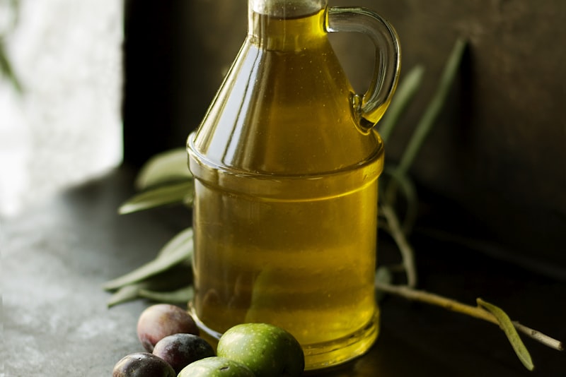 A glass jug of golden olive oil beside a few fresh green and purple olives on a dark surface.
