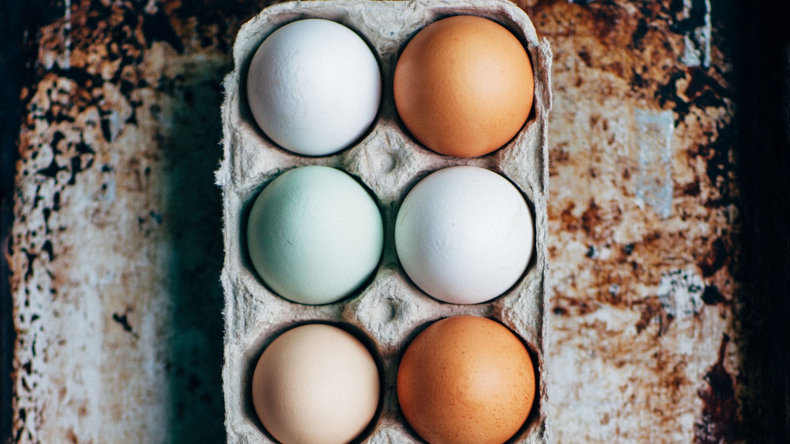 A cardboard carton holding a dozen eggs in shades of white, brown, and pale blue-green.