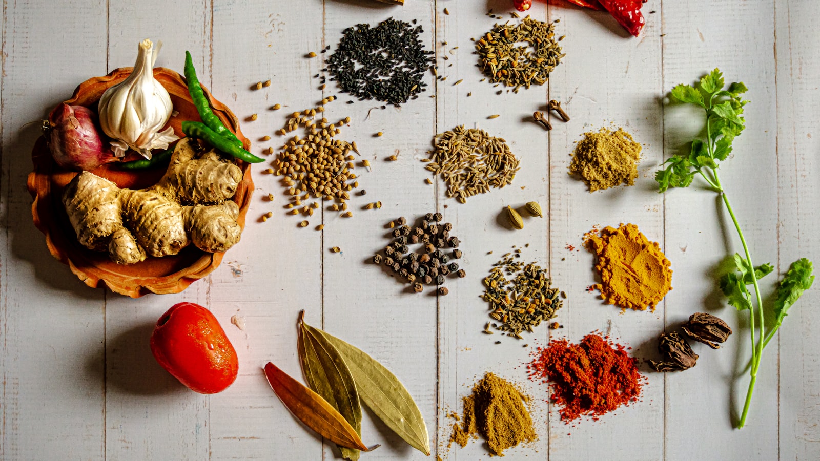 An overhead spread of whole and ground spices, garlic, ginger, chili, and fresh herbs on white wood.