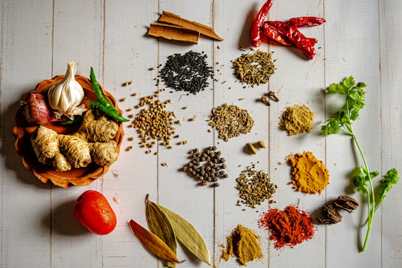 An overhead spread of whole and ground spices, garlic, ginger, chili, and fresh herbs on white wood.