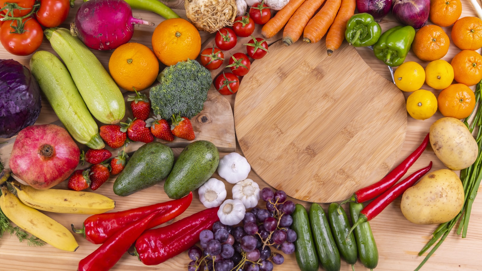 Colorful whole fruits and vegetables arranged in a ring around a round wooden cutting board.