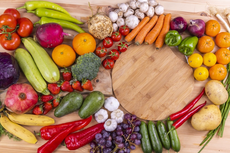 Colorful whole fruits and vegetables arranged in a ring around a round wooden cutting board.